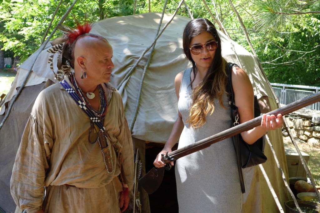 Stephanie Mach (Diné), Curator of North American Ethnographic Collections at the Peabody Museum, in conversation with a reenactor at the Mashantucket Pequot Museum. They are standing in front of a rounded wood and cloth structure, and Mach is holding a musket. Photograph courtesy of Dr. Margaret Bruchac.